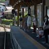 A train station platform with several people waiting, some sitting and some standing, under a wooden canopy The tracks curve to the left, and there are signal lights and a signboard in the background