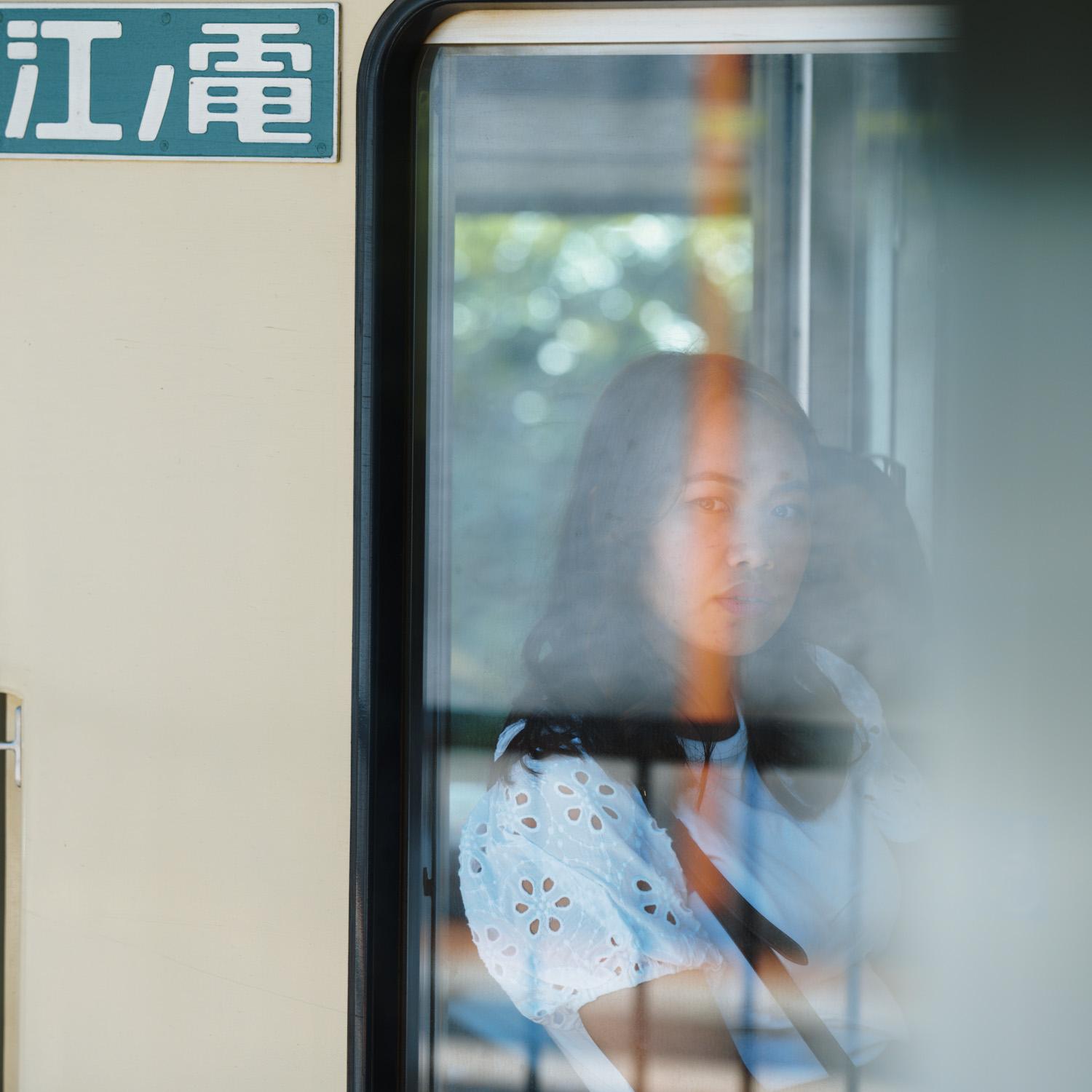 A person with long hair is seen through the window of a train, with a contemplative expression The background outside the train is blurred, and there is a sign with characters in a non-Latin script