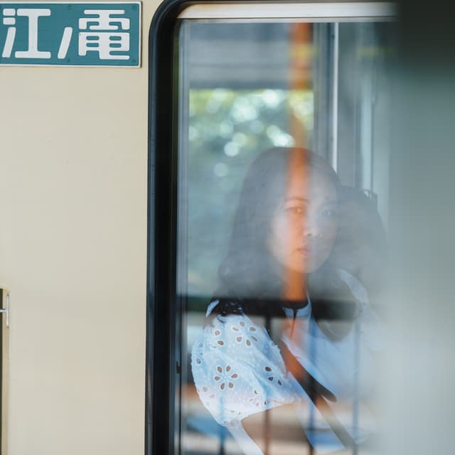 A person with long hair is seen through the window of a train, with a contemplative expression The background outside the train is blurred, and there is a sign with characters in a non-Latin script