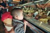 A child is looking at an assortment of gelato flavors in a display case, while someone nearby holds a cone with a scoop of red ice cream