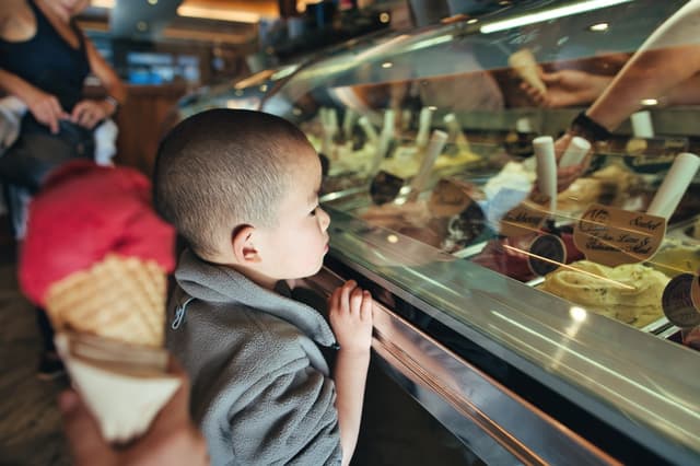 A child is looking at an assortment of gelato flavors in a display case, while someone nearby holds a cone with a scoop of red ice cream